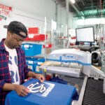 HEATING UP: SquadLocker machine operator Daudi Nabaasa applies a logo to a jersey at a heat-transfer machine at the Warwick company. / PBN PHOTO/DAVE HANSEN