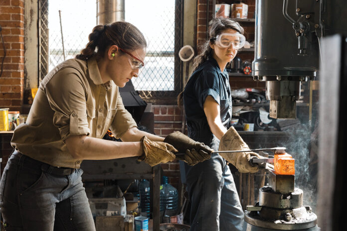 HEATING UP: Alaina Mahoney, left, of Providence-based A.M. Design & Fabrication LLC, works with assistant Erica Compton heating metal in a forge to hammer out a mirror frame for Mahoney’s upcoming product line release.  / COURTESY RUE SAKAYAMA PHOTOGRAPHY