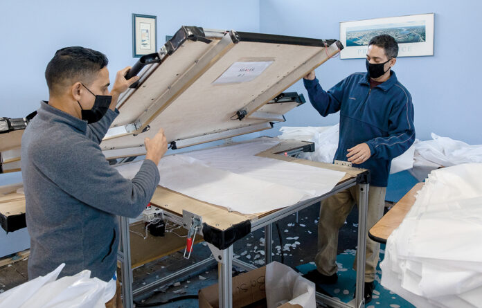 SWITCHING GEARS: Felis Franco, left, and Juan Arrojo load a sealer at Eco Global Manufacturing LLC in Providence. During the COVID-19 pandemic, the product design company switched gears to produce face shields and isolation gowns. / PBN PHOTO/TRACY JENKINS