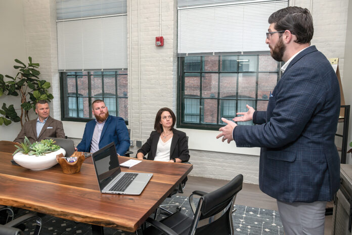 STEADY GROWTH: Cole DeSanty, right, director of client development for The Hire LLC, speaks with his colleagues, from left, CEO Tyler Wentworth; Bryan Soderberg, manager of candidate success; and Chief Operating Officer Erin Pavane. The Providence job placement firm has seen steady revenue growth since offering unlimited PTO from its inception in 2017. / PBN PHOTO/MICHAEL SALERNO
