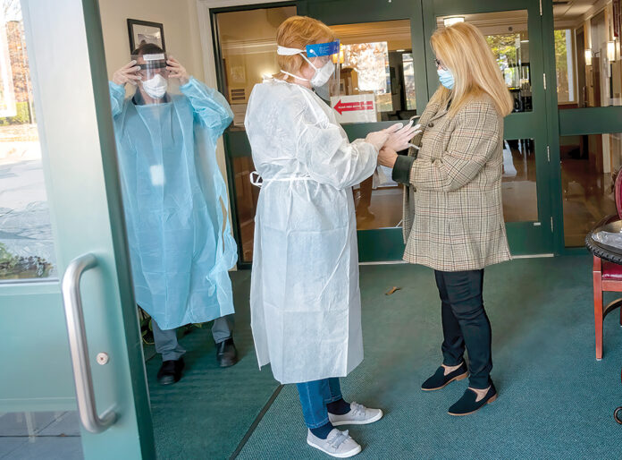 TAKING NO CHANCES: In November 2020, Elizabeth “Libby” Sarro, right, administrator at Bethany Home of Rhode Island, helps office manager Linda Wilson, center, and David Bragga, director of therapy, don protective equipment at the entrance of the Providence nursing home. / PBN FILE PHOTO/MICHAEL SALERNO