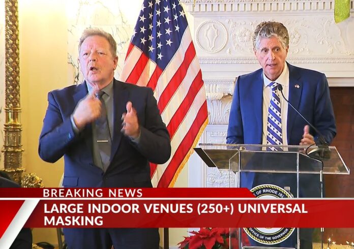 GOV. DANIEL J. MCKEE, right, announces new actions that the state of Rhode Island is taking to slow a surge in COVID-19 cases, at a news briefing at the Statehouse on Wednesday. At left is a sign language interpreter. / SCREENSHOT OF WPRI-TV CBS 12 LIVE STREAM