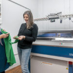 PRACTICAL ­ATTIRE: Propel LLC owner Clare King, left, and Birgit Leitner, product design and development lead, examine an aircraft carrier flight deck jersey Propel manufactures for the U.S. Navy. The company’s whole-garment knitting machine is to the right. / PBN PHOTO/MICHAEL SALERNO