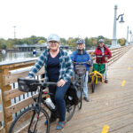 TEMPORARY FIX: Kathleen Gannon, left, board chair of the Rhode Island Bike Coalition, rides across a temporary, boardwalk-style bridge as part of a detour along the East Bay Bike Path going over the Barrington River in Barrington with coalition members Judy Menton, center, and Chris Menton. The old bridge seen in the background is one of two along the bike path that will be replaced following an allocation of federal money for transportation infrastructure improvements awarded to the state in August. PBN PHOTO/KATE WHITNEY LUCEY