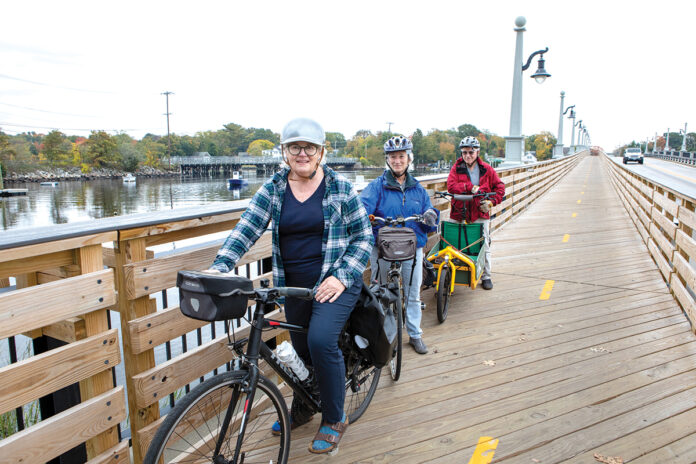 TEMPORARY FIX: Kathleen Gannon, left, board chair of the Rhode Island Bike Coalition, rides across a temporary, boardwalk-style bridge as part of a detour along the East Bay Bike Path going over the Barrington River in Barrington with coalition members Judy Menton, center, and Chris Menton. The old bridge seen in the background is one of two along the bike path that will be replaced following an allocation of federal money for transportation infrastructure improvements awarded to the state in August. PBN PHOTO/KATE WHITNEY LUCEY