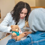 ON THE MEND: Anna Rezk, a Warren Alpert Medical School student, checks the casting work of a youngster who was assigned to treat a teddy bear with a broken arm at the Black Men in White Coats youth summit at the Brown University medical school in October.  PBN PHOTO/MICHAEL SALERNO