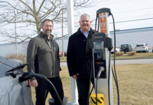 AMPED UP: Zachary Cobb, left, Hexagon Manufacturing Intelligence vice president of services, and Steven Ilmrud, vice president of operations, demonstrate one of the charging stations at the North Kingstown company that are part of the company’s ambitious plans to offset the emissions it produces with renewable energy or credits by 2030.  PBN PHOTO/ELIZABETH GRAHAM