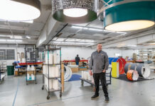 SEEING THE LIGHT: Ian Prichett, CEO and president of Lumetta Inc., stands on the floor of the Warwick lighting manufacturer with some of its designs hanging overhead. He says technologies and product designs evolve quickly in the lighting world, so he’s constantly spending time on the floor and learning from each ­department. PBN PHOTO/MICHAEL SALERNO
